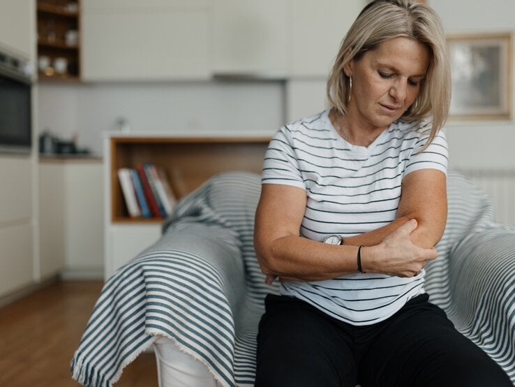 Frau sitzt auf Bett und greift sich an den Ellenbogen.