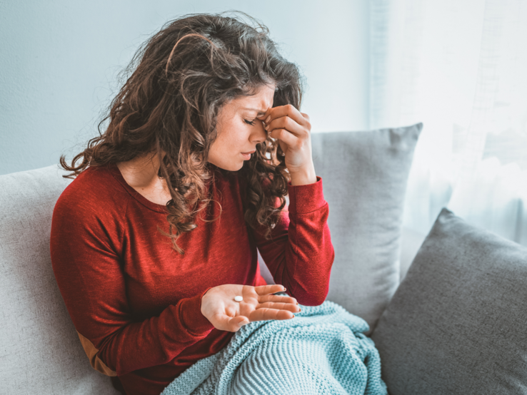 Junge Frau mit langen braunen Locken sitzt erschöpft auf dem Sofa, hält Tabletten in der Hand und reibt sich die Stirn.