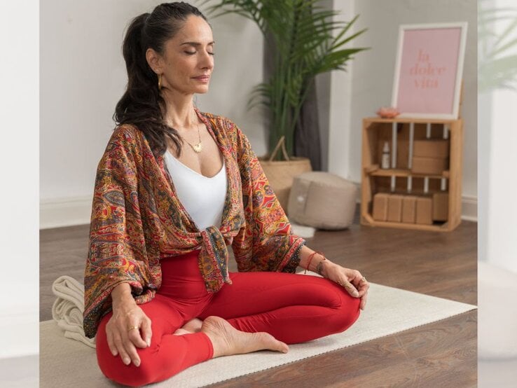 Eine Person sitzt in einer entspannten Meditationshaltung, gekleidet in einem gemusterten Kimono und roten Leggings. Der Hintergrund zeigt eine Zimmerpflanze, eine Holzablage, in der Kissen und dekorative Gegenstände stehen, sowie einen Schriftzug "la dolce vita". | © YogaEasy GmbH