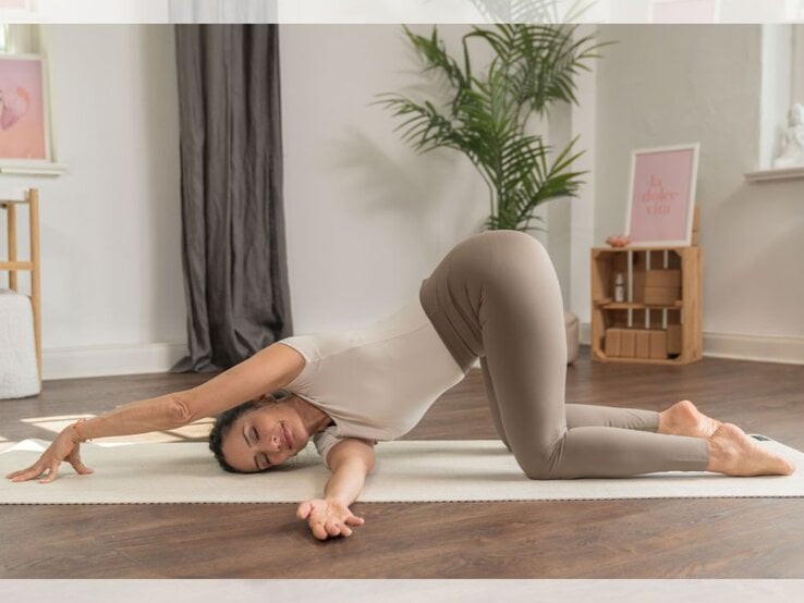 Eine Frau führt eine Yoga-Dehnübung aus, bei der sie mit einem Arm gestreckt auf einer Matte liegt, während sie den anderen Arm nach vorne ausstreckt. Im Hintergrund sind ein Zimmerpflanze, eine braune Holzablage und Wanddekorationen sichtbar. | © YogaEasy GmbH