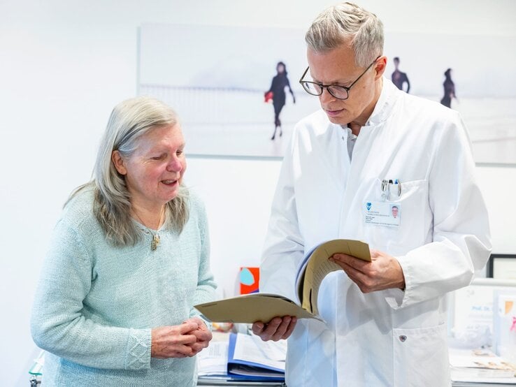 Ältere Frau im hellblauen Pullover spricht lächelnd mit einem grauhaarigen Arzt im weißen Kittel, der ein Dokument durchblättert.