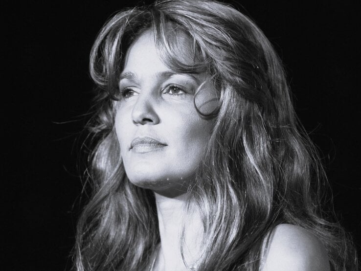 Porträt einer nachdenklich blickenden Frau mit langen, gewellten Haaren vor schwarzem Hintergrund im Scheinwerferlicht.