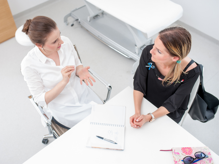 Ärztin im Gespräch mit einer Patientin, beide sitzen an einem Tisch, Perspektive von oben.