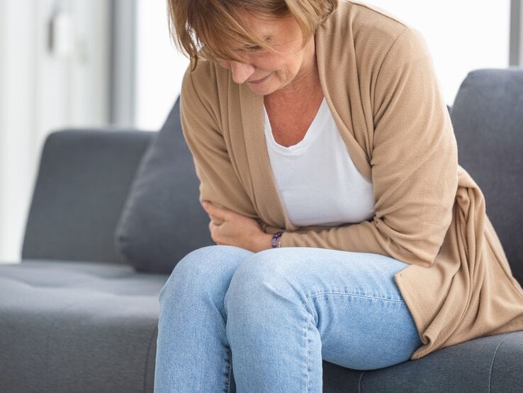 Eine Frau hält sich vor Schmerzen den Bauch und sitzt dabei auf einem grauen Sofa.