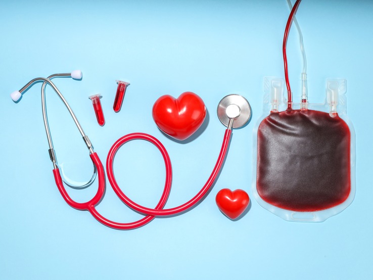 Medizinisches Stethoskop, ein Blutbeutel, zwei Blutprobenröhrchen und zwei rote Herzmodelle auf blauem Hintergrund arrangiert.