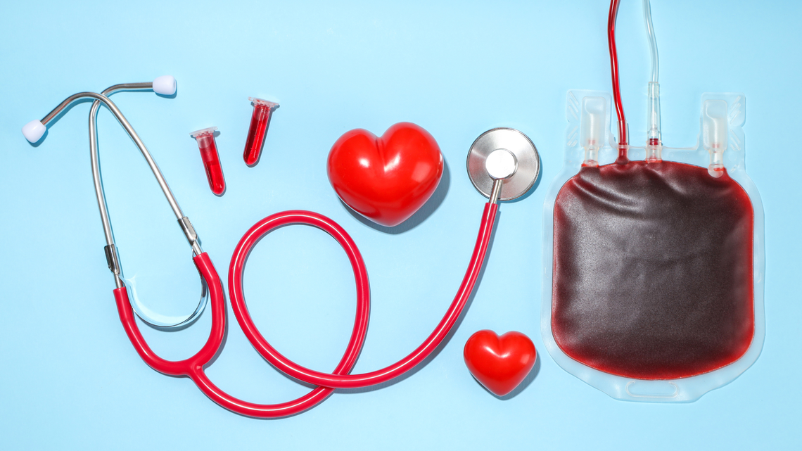 Medizinisches Stethoskop, ein Blutbeutel, zwei Blutprobenröhrchen und zwei rote Herzmodelle auf blauem Hintergrund arrangiert.