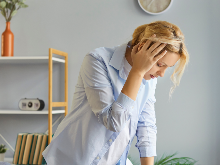 Eine blonde Frau in hellem Hemd stützt sich erschöpft auf einen Tisch in einem modernen Raum mit Holzregalen und Dekoration.