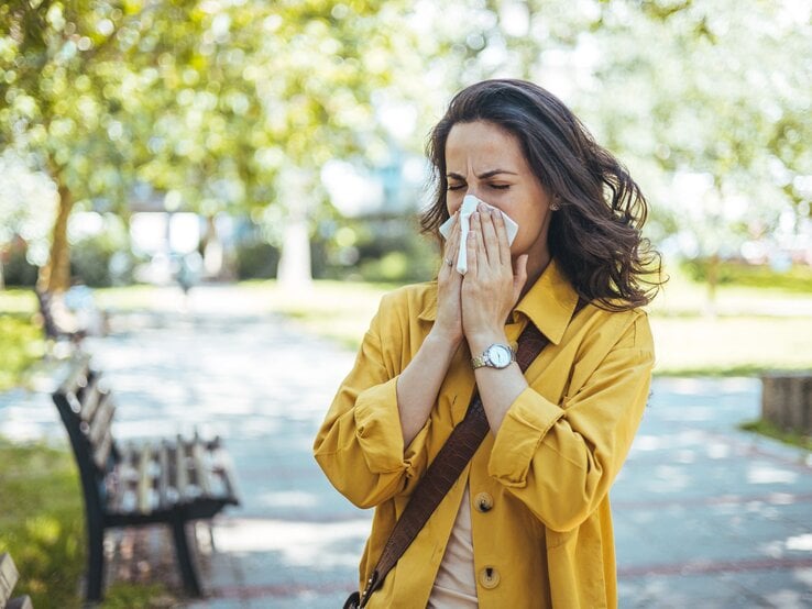Eine Frau mit dunklen Haaren und gelbem Mantel niest aufgrund von Heuschnupfen  mit geschlossenen Augen in ein Taschentuch in einem sonnigen Park.