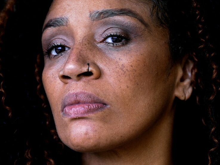 Eine Frau mit dunkler Haut, Nasenpiercing und offenen Locken blickt ernst in die Kamera, ihr Gesicht zeigt Sommersprossen.