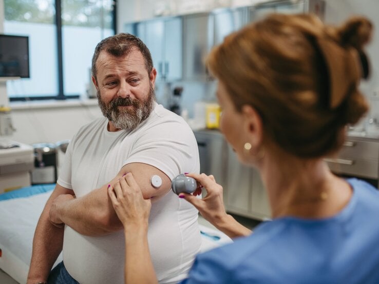 Älterer Mann mit grauem Bart lässt in Klinik von Ärztin im blauen Kittel seinen Glukosesensor am Oberarm scannen.