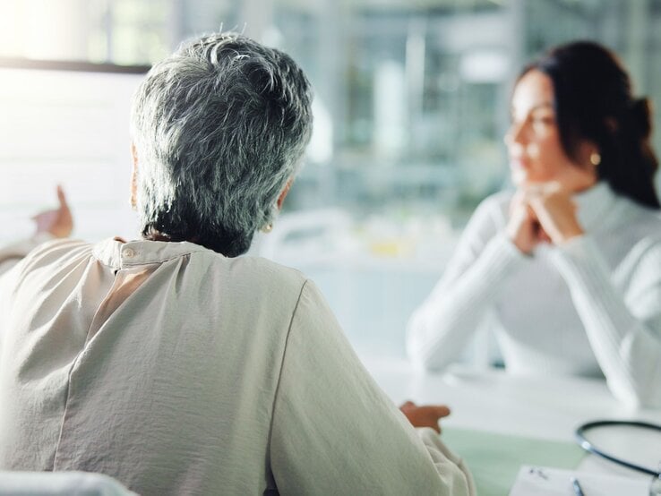 Eine ältere Frau mit grazem Kurzhaarschnitt spricht ernst mit einer jungen Frau in weißem Rollkragenpullover am Schreibtisch.
