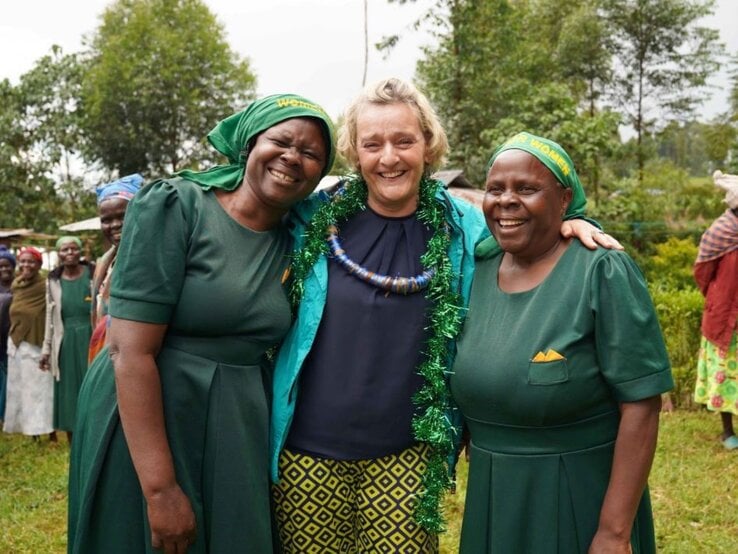 Glückliche Szene: Drei Frauen umarmen sich im Grünen, lachen in die Kamera, im Hintergrund weitere Frauen in traditioneller Kleidung.