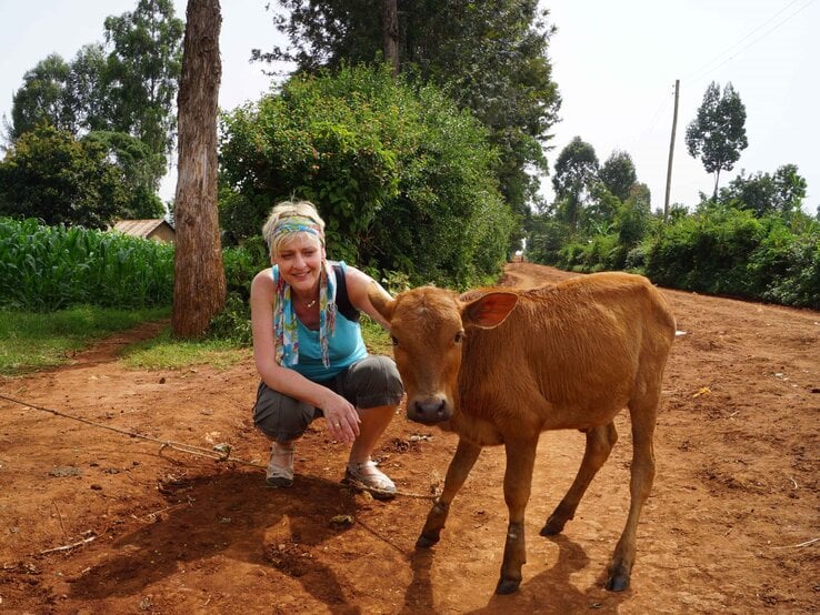Eine lächelnde Frau mit bunter Haarbinde hockt neben einem braunen Kalb auf einem roten Sandweg vor dichtem Grün. | © GOLDENE BILD der FRAU