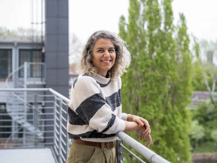 Junge Frau mit grauen Locken und gestreiftem Pullover lehnt lächelnd am Balkongeländer vor grünen Bäumen und moderner Architektur.