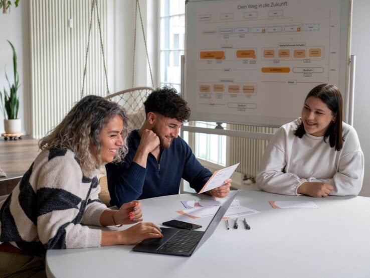 Drei junge Erwachsene sitzen lachend an einem Konferenztisch, vor ihnen Laptop, Karten und Stifte, im Hintergrund ein Flipchart. | © GOLDENE BILD der FRAU