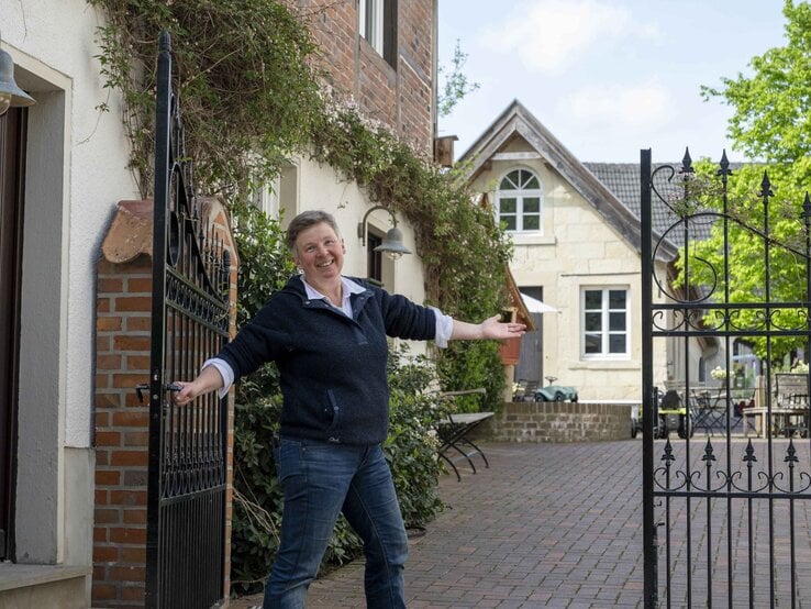 Freundlich lachende Frau mittleren Alters mit kurzem Haar öffnet ein schwarzes Tor zu einem idyllischen Innenhof mit Landhausflair.