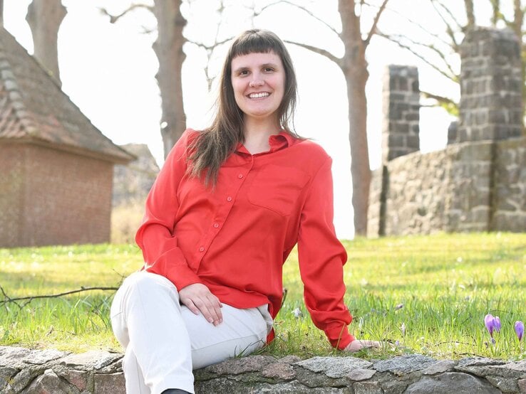 Junge Frau mit braunen Haaren, roter Bluse und weißer Hose sitzt lächelnd auf einer Mauer vor alten Steinruinen im Sonnenschein.