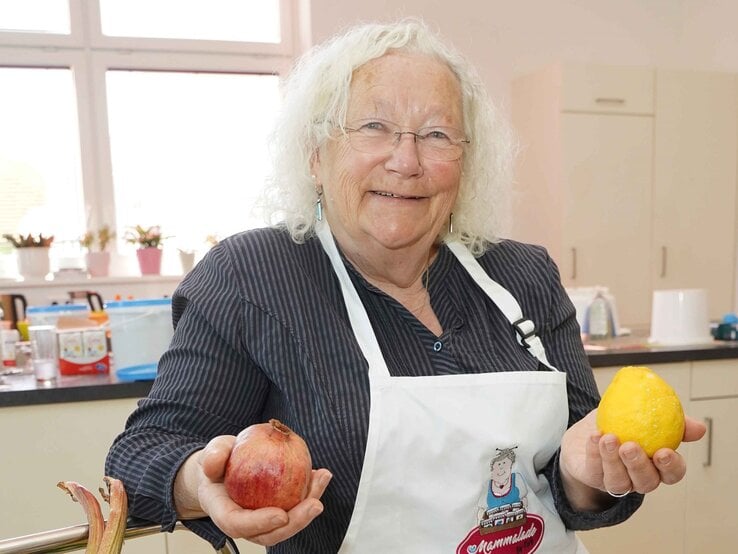 Ältere Frau mit weißen Locken und Brille lächelt in Küche, hält Granatapfel und Zitrone, trägt gestreifte Bluse und Schürze.