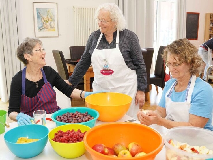 Drei ältere Frauen bereiten gemeinsam in heller Küche Obst vor, lachen dabei herzlich, umgeben von bunten Schüsseln. | © GOLDENE BILD der FRAU