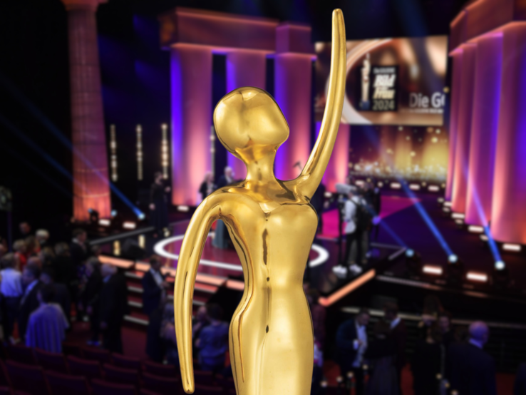 GOLDENE BILD der FRAU Statue mit erhobenem Arm vor beleuchteter Bühne und Publikum bei festlicher Gala mit lila Lichtakzenten.