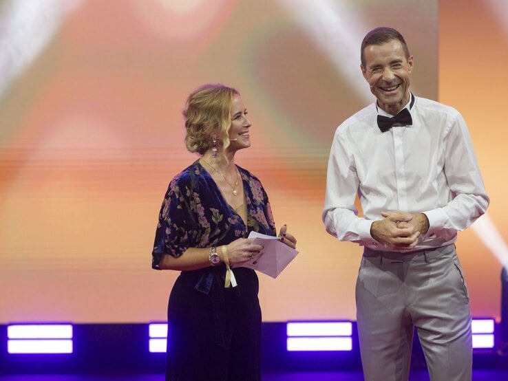 Eine blonde Frau in gemusterter Bluse und ein Mann im weißen Hemd mit Fliege lachen auf einer Bühne vor orangefarbenem Hintergrund. | © Guido Ohlenbostel