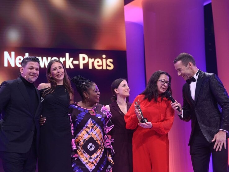 Eine lächelnde Gruppe aus drei Frauen und einem Mann posiert mit einer Preisträgerin in rotem Kleid, auf einer Bühne mit violettem Licht. | © Henning Scheffer Photography 