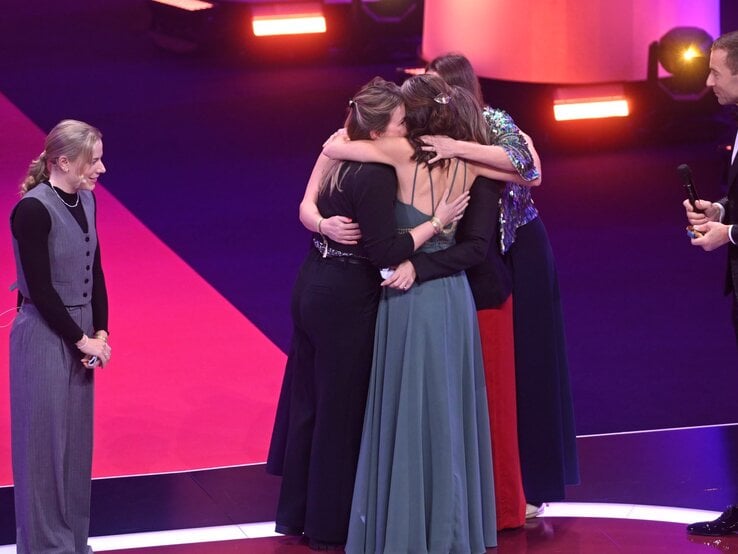 Vier Frauen umarmen sich emotional auf einer Bühne mit pinkem Teppich, während ein Mann im Smoking mit Mikrofon lächelnd daneben steht. | © Henning Scheffer Photography 
