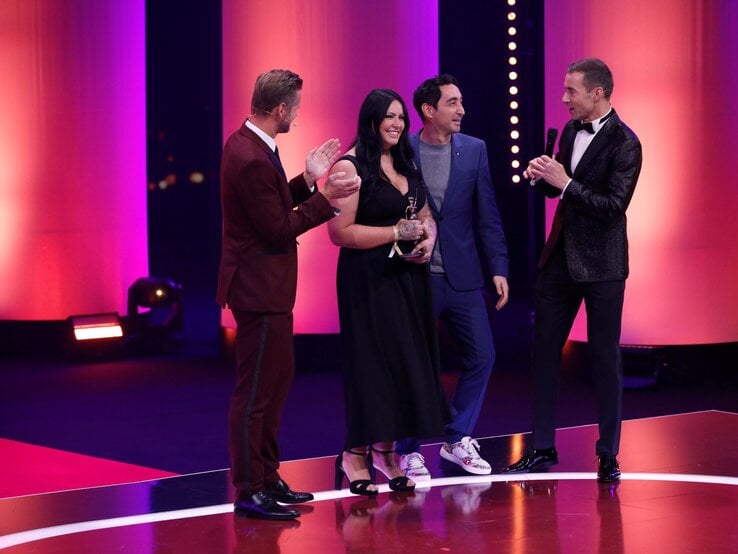 Eine Frau mit langen schwarzen Haaren und schwarzem Kleid hält eine Trophäe, während drei Männer in Anzügen auf einer beleuchteten Bühne applaudieren. | © Henning Scheffer Photography 