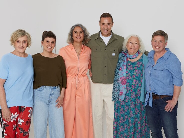 Fünf Frauen und ein Mann unterschiedlichen Alters stehen lächelnd nebeneinander vor weißem Hintergrund und wirken entspannt.