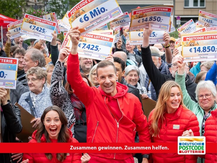 Gruppe von Gewinnern der Deutschen Postcode Lotterie hält lachend ihre Gewinnschecks in die Höhe.