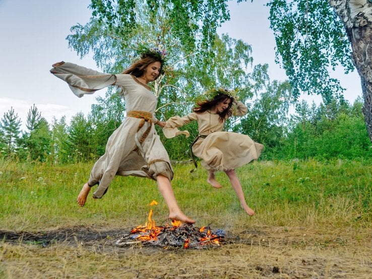 Zwei Frauen mit Blumenkränzen springen über ein Sonnenwend-Feuer.