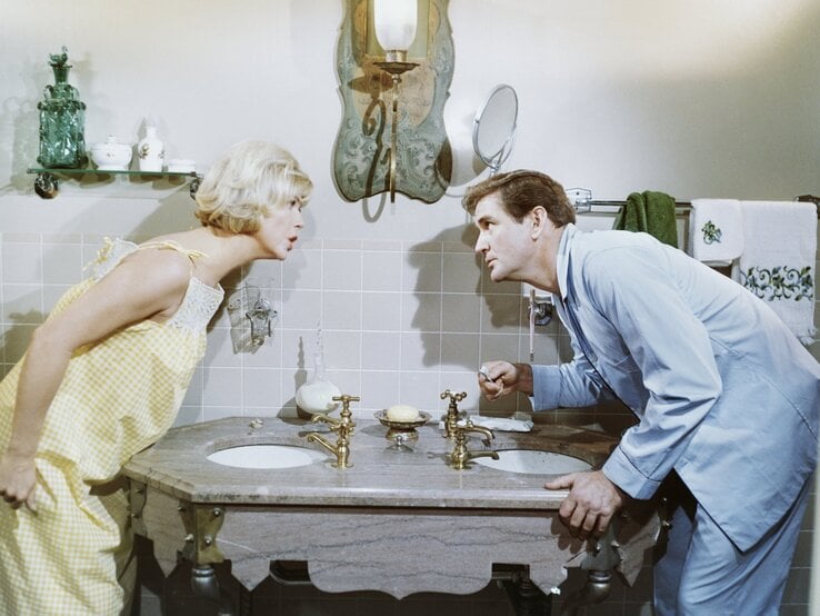 Ein Mann und eine Frau in Schlafkleidung beugen sich gleichzeitig über ein Doppelwaschbecken im Badezimmer und schauen sich mit ernsten, fast konfrontativen Blicken an. Die Frau trägt ein gelb kariertes Nachthemd, der Mann einen hellblauen Pyjama. Das Badezimmer hat ein Vintage-Ambiente mit Messingarmaturen, einem Spiegel mit geschwungenem Rahmen und handbestickten Handtüchern.