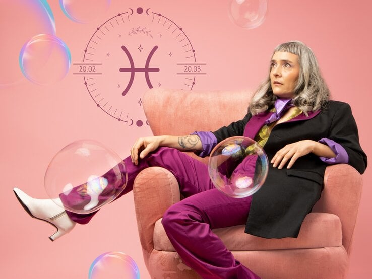 Eine Frau mit langen grauen Haaren trägt einen lila Retro-Anzug und sitzt in einem Sessel, das Bein lässig über die Lehne geschlagen. Sie ist umgeben von Seifenblasen und neben ihr ist das astrologische Symbol des Sternzeichens Fische in einem Kreis zu sehen.