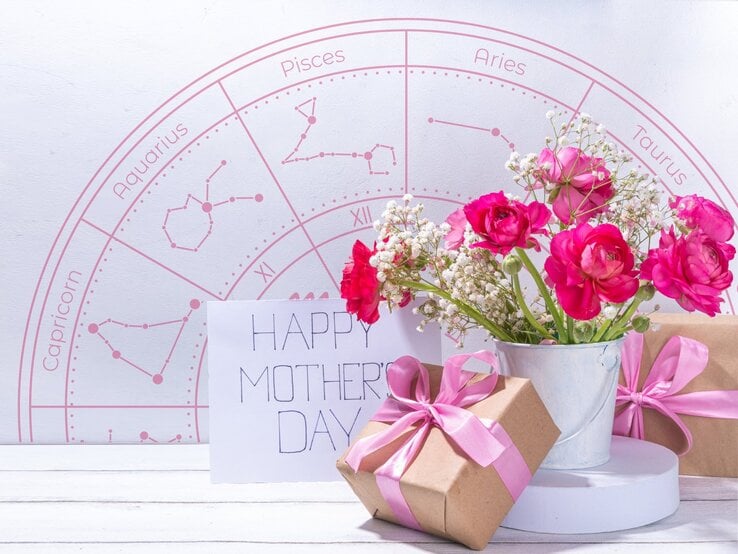 Ein liebevoll arrangiertes Muttertagsstillleben mit pinken Ranunkeln und Schleierkraut in einem weißen Metalltopf, daneben zwei in braunes Papier eingepackte Geschenke mit rosa Schleifen und eine Karte mit der Aufschrift ‚Happy Mother’s Day‘. Im Hintergrund ist ein rosafarbener Tierkreis mit Sternzeichen wie Pisces, Taurus und Capricorn zu sehen – eine harmonische Verbindung aus Astrologie, Blumen und Muttertagsliebe.