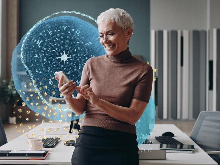 Geschäftsfrau mit grauen Haaren, die ein Smartphone benutzt und lächelt, während sie sich an den Schreibtisch lehnt.