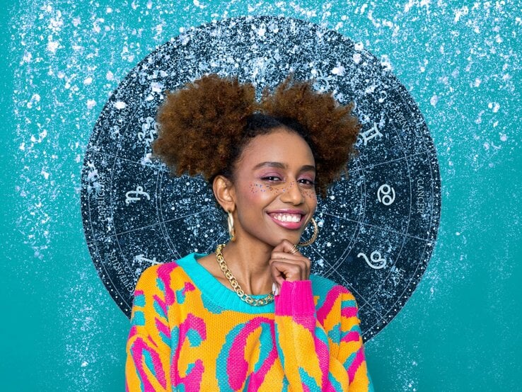 Eine fröhliche Frau mit lockigem Haar steht vor einem blauen Hintergrund mit einem großen astrologischen Rad. Sie trägt einen bunten Pullover mit leuchtenden Farben und lächelt in die Kamera. Auf ihrem Gesicht sind glitzernde Punkte zu sehen, die ihre Wangen und Nase verzieren.