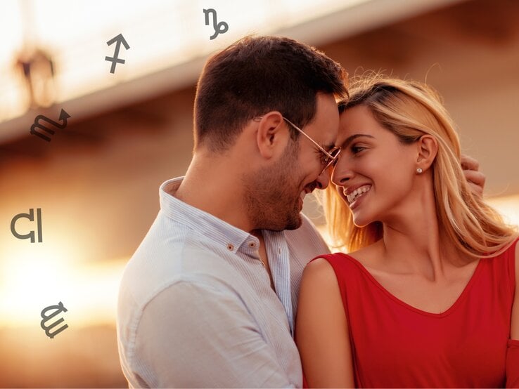 Ein lächelndes Paar steht einander nahe und genießt einen romantischen Moment im Sonnenuntergang. Der Mann trägt eine Brille und ein helles Hemd, während die Frau ein rotes Kleid trägt. Sie berühren sanft ihre Stirnen und strahlen Freude und Zuneigung aus. Im Hintergrund sind astrologische Symbole zu sehen, was eine symbolische und astrologische Bedeutung suggeriert. Die warme Beleuchtung und der unscharfe Hintergrund verstärken die intime und liebevolle Atmosphäre.