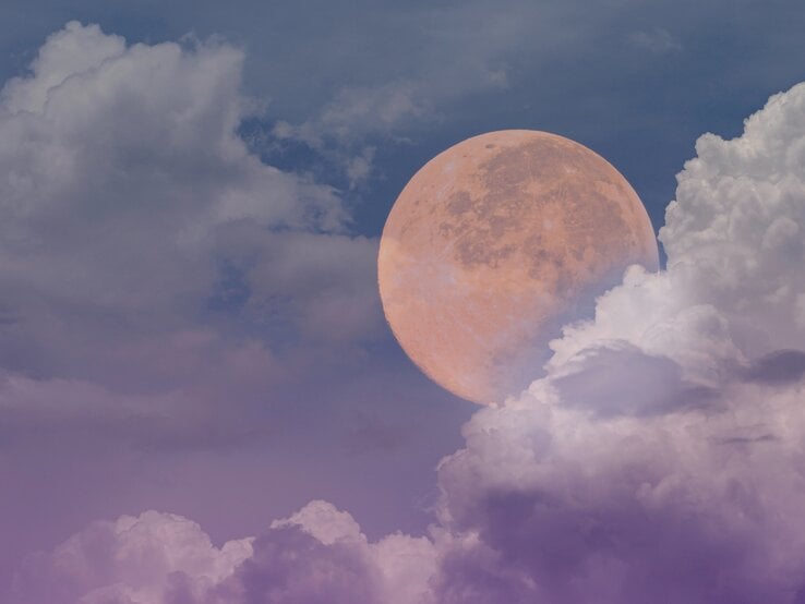 Ein großer, leuchtender Vollmond scheint durch eine teilweise bewölkte Himmelsszene. Der Himmel ist in verschiedenen Blautönen gefärbt, die von einem tiefen, dunklen Blau bis hin zu einem sanften Lila reichen. Die Wolken sind weich und flauschig, in hellen Weiß- und Lilatönen, und umrahmen den Mond, der in warmen, orangefarbenen Tönen leuchtet. Die Szenerie vermittelt eine ruhige und mystische Atmosphäre.