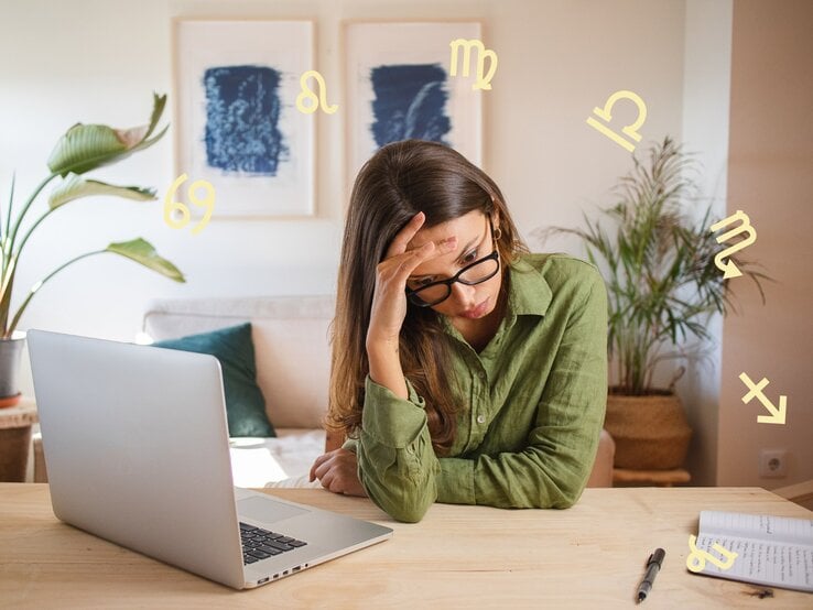 Das Bild zeigt eine Frau, die vor einem Laptop an einem Schreibtisch sitzt und erschöpft oder gestresst wirkt. Sie stützt ihren Kopf mit einer Hand und trägt eine Brille sowie ein grünes Hemd. Auf dem Tisch liegen ein Notizbuch und ein Stift. Im Hintergrund sind Zimmerpflanzen und zwei gerahmte Bilder an der Wand zu sehen. Um sie herum schweben verschiedene astrologische Symbole, was auf einen Zusammenhang mit Astrologie oder Sternzeichen hinweist. Die Szene vermittelt eine Atmosphäre von Müdigkeit oder Überforderung in einer häuslichen oder Arbeitsumgebung.