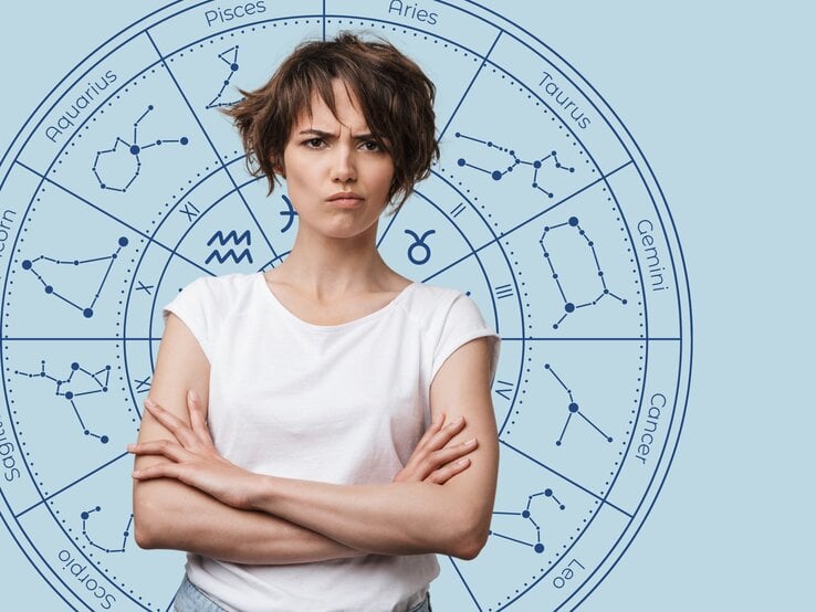 Das Bild zeigt eine junge Frau mit kurzen, braunen Haaren und einem verärgerten Gesichtsausdruck. Sie trägt ein weißes T-Shirt und hat die Arme verschränkt. Im Hintergrund ist ein astrologisches Rad mit den Sternzeichen und deren Konstellationen auf einem hellblauen Hintergrund abgebildet.