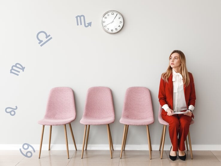 Das Bild zeigt eine junge Frau in einem eleganten, roten Hosenanzug und weißer Bluse, die auf einem rosa Stuhl sitzt und nachdenklich zur Seite blickt. Sie sitzt in einer Reihe von vier rosa Stühlen vor einer hellgrauen Wand. An der Wand hängen astrologische Symbole der Sternzeichen, die scheinbar um eine Uhr herum angeordnet sind.