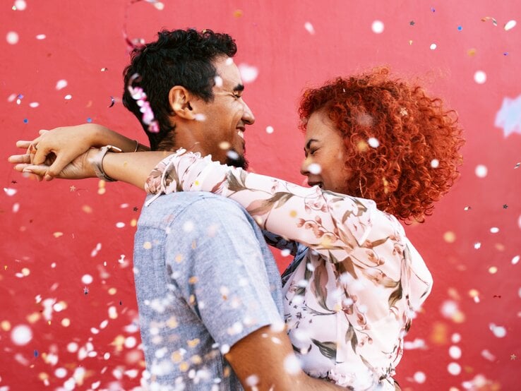 Das Bild zeigt ein glückliches Paar, das sich umarmt und in die Augen schaut. Die Frau hat leuchtend rote, lockige Haare und trägt eine Bluse mit Blumenmuster. Der Mann hat dunkle, lockige Haare und trägt ein hellblaues Hemd. Beide lächeln und scheinen sich sehr zu freuen. Im Hintergrund ist eine rote Wand zu sehen, und über dem Paar fliegt bunter Konfetti, was eine fröhliche und feierliche Atmosphäre erzeugt.