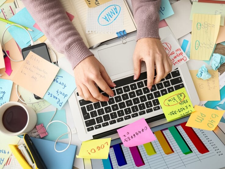 Eine Person tippt auf der Tastatur eines Laptops, umgeben von einem chaotischen Schreibtisch mit zahlreichen Haftnotizen, Dokumenten, einem Smartphone, einem Kaffeebecher und farbigen Diagrammen. Der Arbeitsplatz wirkt unorganisiert, mit Kabeln und zerknülltem Papier, was auf eine stressige oder überladene Arbeitssituation hinweist.