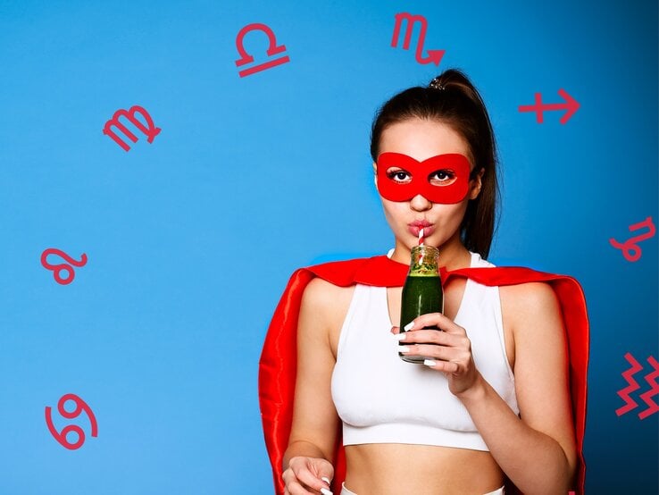 ine junge Frau trägt eine rote Superheldenmaske und ein passendes rotes Cape, während sie einen grünen Smoothie aus einer Flasche mit einem Strohhalm trinkt. Sie trägt ein weißes Sport-Top und steht vor einem blauen Hintergrund, umgeben von roten astrologischen Symbolen verschiedener Sternzeichen.