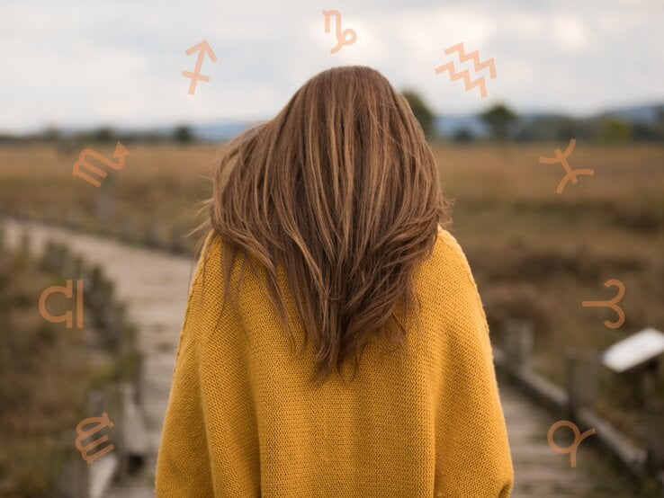 Eine Frau mit langen braunen Haaren in einem gelben Pullover steht auf einem Holzsteg in einer weiten Landschaft. Um ihren Kopf herum sind astrologische Symbole verschiedener Sternzeichen angeordnet.