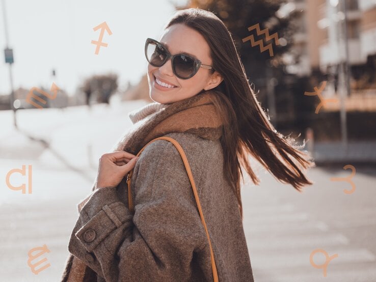 Eine lächelnde Frau trägt eine Sonnenbrille und einen dicken Mantel, während sie sich im Freien umdreht und in die Kamera schaut. Um sie herum sind Symbole der Tierkreiszeichen schwebend zu sehen. Der Hintergrund ist eine städtische Umgebung mit verschwommenen Gebäuden und Bäumen, was eine herbstliche, entspannte Atmosphäre vermittelt.