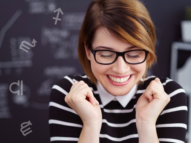 Eine lächelnde Frau mit Brille und gestreiftem Oberteil zeigt eine freudige Reaktion, während sie die Augen geschlossen und die Fäuste leicht geballt hat. Im Hintergrund ist eine Tafel mit verschwommenen, handgeschriebenen Notizen zu sehen. Um sie herum schweben verschiedene Symbole der Tierkreiszeichen, was auf eine astrologische Verbindung oder ein thematisches Setting hinweist. Die Szene vermittelt positive Emotionen, Erfolg oder einen glücklichen Moment.