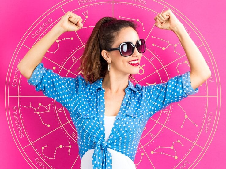 Eine Frau in einer blauen, gepunkteten Bluse posiert selbstbewusst vor einem astrologischen Rad, das verschiedene Sternzeichen zeigt. Sie trägt eine Sonnenbrille und hebt ihre Arme in einer kraftvollen Pose. Das Bild vermittelt Optimismus, Selbstbewusstsein und Erfolg.