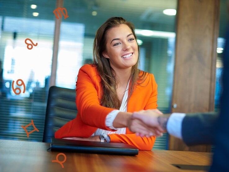 Eine lächelnde Frau in einem orangefarbenen Blazer sitzt in einem modernen Büro und schüttelt die Hand einer anderen Person, die teilweise im Bild sichtbar ist. Im Hintergrund schweben astrologische Symbole wie Löwe, Jungfrau, Krebs und Zwillinge in der Luft.