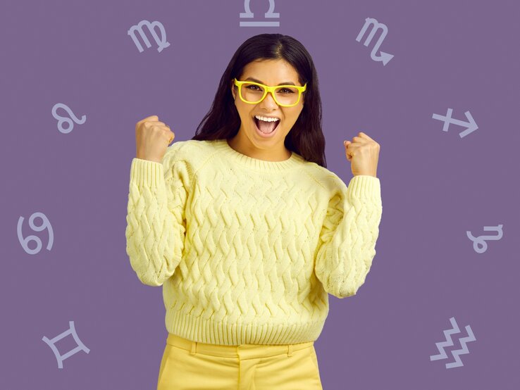 Eine fröhliche Frau mit langen, dunklen Haaren, die gelbe Brille und einen gelben Pullover trägt, zeigt freudig beide Fäuste. Im Hintergrund sind astrologische Symbole auf einem lilafarbenen Hintergrund verteilt.
